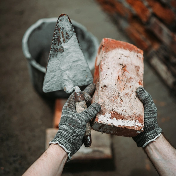 Nahaufnahme von Händen, die Ziegel und Kelle mit nassem Zement halten.